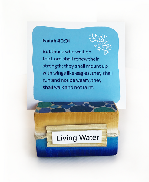 Image shows sea-inspired scripture card holder holding a sky blue color scripture card found within the sea-inspired scripture card set. “Living Water” label in front of holder reminds us of our need to quench our thirst with spiritual truth. Groove designed to stand up multiple cards at one time. Playful circular pattern on top creatively displays pebbles on the sea floor. Seaside twine wraps around outer border.  Hand-painted ocean blues meet sandy beach tones along all sides.   tribegift.com