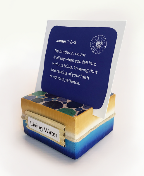 Image shows sea-inspired scripture card holder holding a deep blue sea color scripture card found within the sea-inspired scripture card set. “Living Water” label in front of holder reminds us of our need to quench our thirst with spiritual truth. Groove designed to stand up multiple cards at one time. Playful circular pattern on top creatively displays pebbles on the sea floor. Seaside twine wraps around outer border.  Hand-painted ocean blues meet sandy beach tones along all sides.   tribegift.com